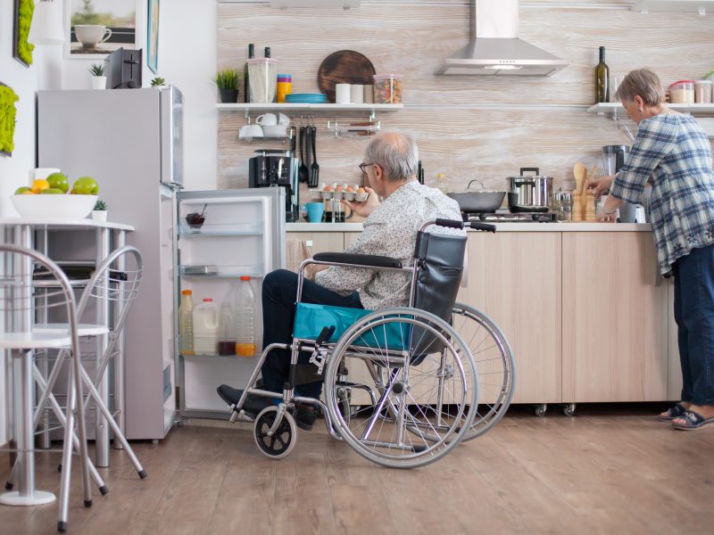 Handicapped man helping wife at the kitchen by taking eggs carton from refrigerator. Senior woman helping handicapped husband. Living with disabled person with walking disabilities